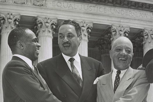 Thurgood Marshall stands in front of the Supreme Court of the United States with George Edward Chalmer Hayes and James Nabrit. The three men are smiling and shaking hands.