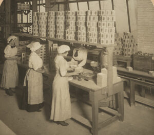 Five workers dressed in long skirts and hair coverings stand on both sides of a table - three on the left, two on the right. Each is packing sugar.